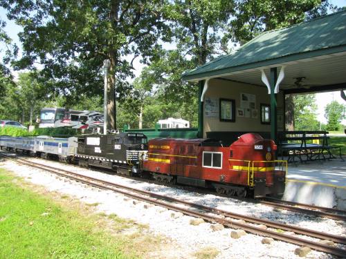 A miniature train with a red engine and several cars is parked at a small station surrounded by trees.