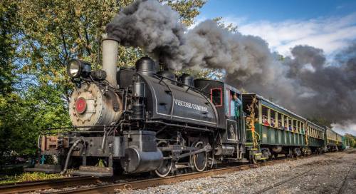 A vintage steam locomotive emits smoke while pulling a green passenger car along a scenic railway.