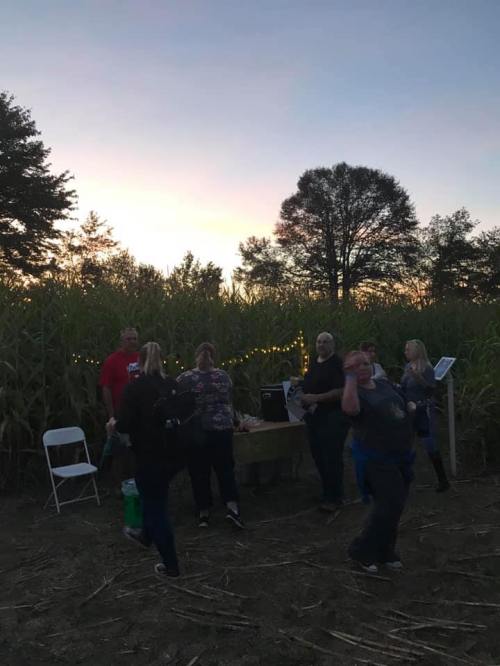 A group of people gathered in a cornfield at sunset, with lights and a table set up for an outdoor event.