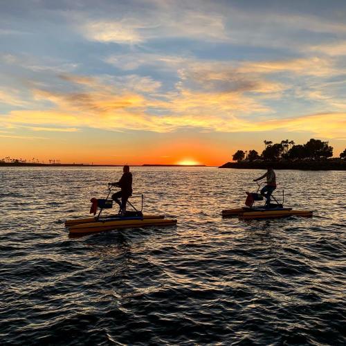 Two people ride pedal boats on a calm waterway at sunset, with vibrant orange and blue skies in the background.