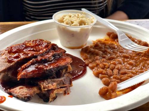 A plate with sliced barbecue brisket, baked beans, and a small cup of macaroni salad.