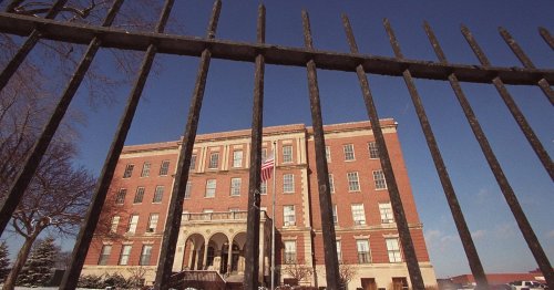 A historic brick building behind a black wrought iron fence, with an American flag visible in front.