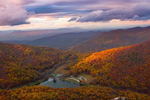 A scenic view of colorful autumn foliage over rolling mountains and a serene lake under a dramatic sky.