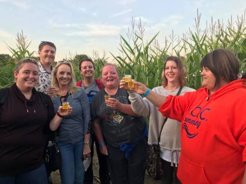 A group of eight people smiling and holding drinks, standing in a cornfield during a gathering or celebration.