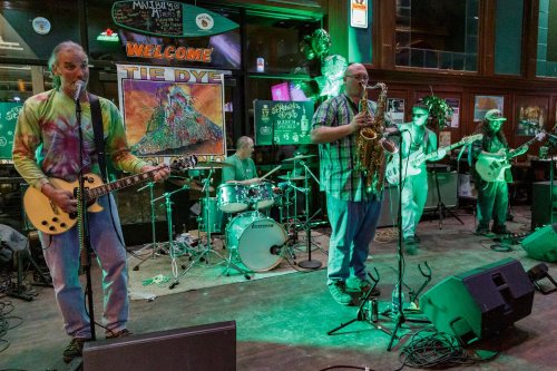 A lively band performs on stage in a bar, featuring guitars, drums, and a saxophone, with colorful lighting.