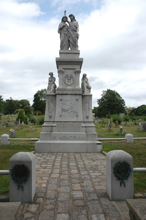 A tall monument with two statues atop, surrounded by a cemetery with gravestones and greenery in the background.