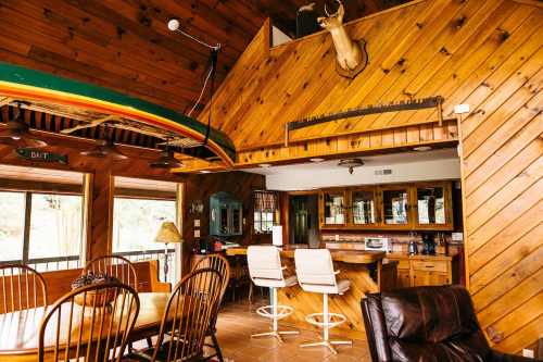 Cozy wooden interior with a dining area, bar stools, and a mounted deer head, featuring a kayak on the ceiling.