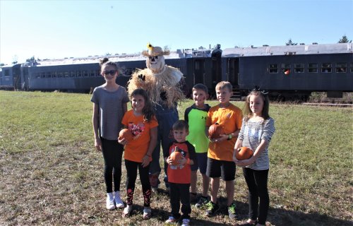 A group of six children poses with a scarecrow and pumpkins near a train in a sunny field.