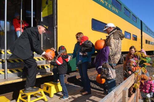 A conductor hands pumpkins to families boarding a train on a sunny day, with colorful decorations nearby.