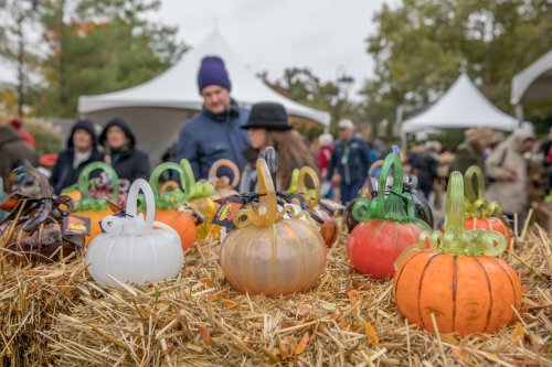Colorful glass pumpkins displayed on hay bales, with people browsing in the background at an outdoor event.