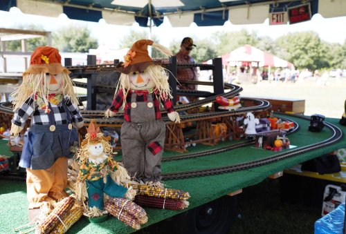 Three scarecrows in plaid outfits stand near a model train setup at an outdoor fair, with colorful decorations around.