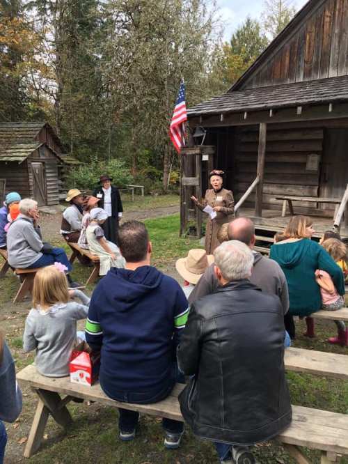 A group of people seated outdoors, listening to a speaker in period costume near a log cabin with an American flag.