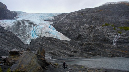 A person stands near a glacial landscape with rocky terrain and a melting glacier in the background.