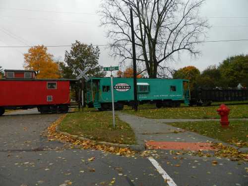 A red and teal train car at a railroad crossing, surrounded by autumn leaves and a tree in a cloudy sky.