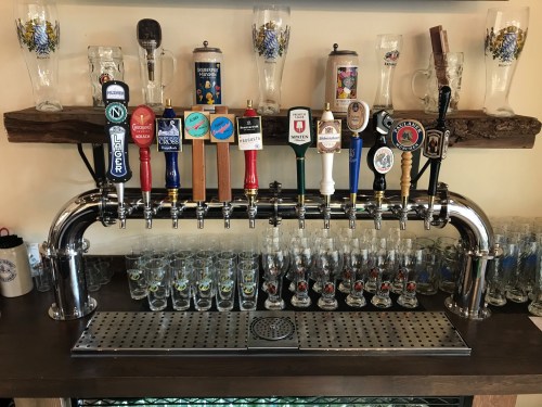 A shiny beer tap with multiple handles, set against a wooden shelf with glasses and a bar counter below.