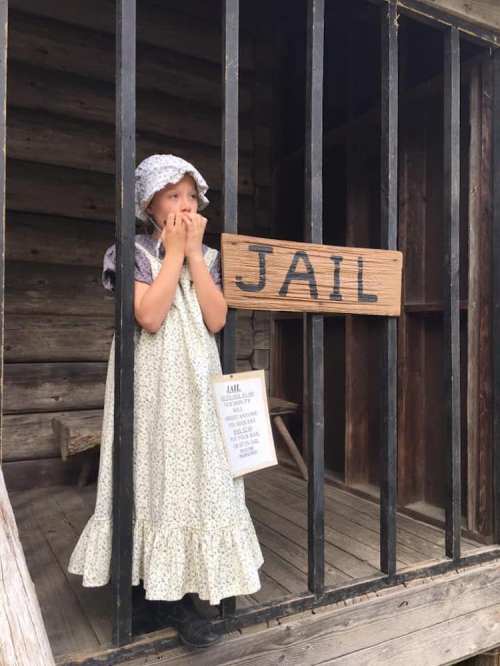 A young girl in a vintage dress stands behind bars in a wooden jail, looking surprised, with a sign labeled "JAIL."