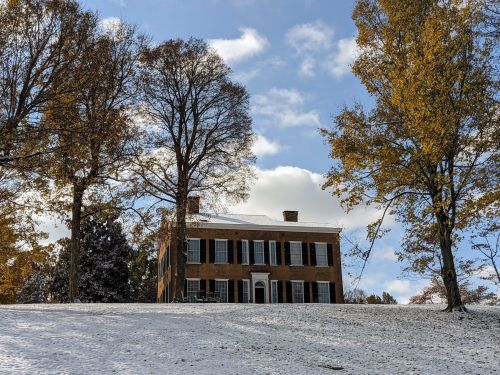 A historic brick house on a snowy hill, surrounded by tall trees with autumn leaves under a blue sky.