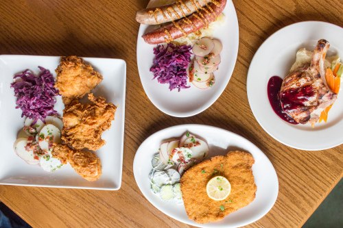 A table with four plates of food: fried chicken, sausages, pork chop, and schnitzel, accompanied by sides and garnishes.