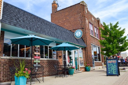 A charming brick storefront with outdoor seating and umbrellas, featuring colorful signage and potted plants.