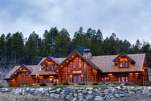 A large log cabin with warm lights, surrounded by trees and a stone wall, under a cloudy sky.