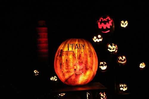 A carved pumpkin featuring an Italian scene, illuminated among other glowing jack-o'-lanterns in a dark setting.