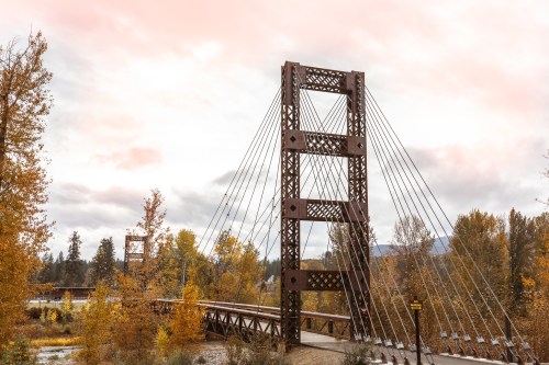 A suspension bridge with a metal frame spans a river, surrounded by autumn trees and a cloudy sky.
