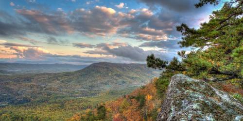 A panoramic view of rolling hills and valleys under a colorful sky, with trees in the foreground and distant mountains.