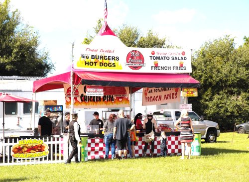 A colorful food stand with a red and white checkered table, offering burgers, hot dogs, fries, and tomato salad.