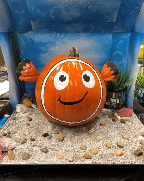 A painted pumpkin resembling a clownfish, with a smiling face and fins, displayed on a sandy surface with pebbles.