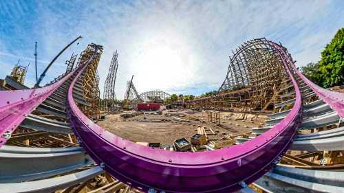 A panoramic view of a wooden roller coaster under construction, with tracks and supports visible against a bright sky.