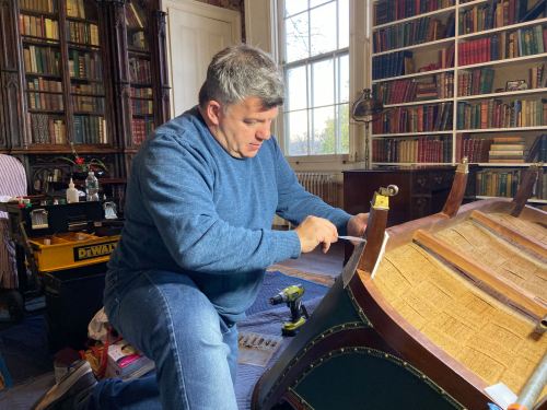 A man in a blue sweater works on a wooden boat in a room filled with books and tools.