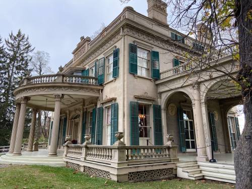 A historic mansion with green shutters, columns, and a wraparound porch, surrounded by trees and a cloudy sky.