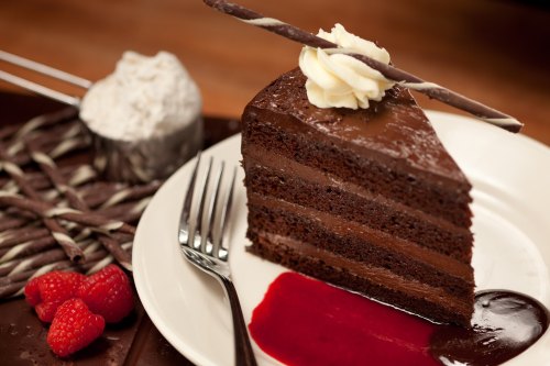 A slice of rich chocolate cake with layers, topped with cream and a chocolate stick, served with raspberry sauce and berries.