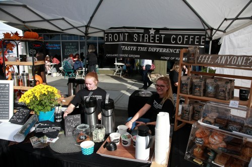 Two baristas serve coffee at a market stall, with a sign reading "Front Street Coffee" and various coffee products displayed.