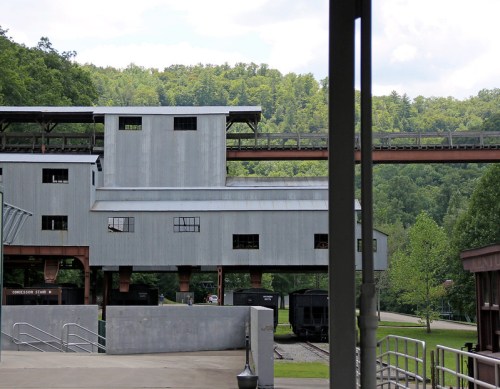 A gray industrial building with a conveyor belt, surrounded by green trees and a clear sky.