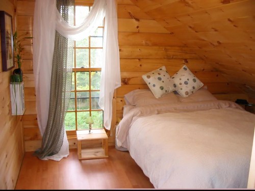 Cozy wooden bedroom with a bed, decorative pillows, a small table, and a window draped with sheer curtains.