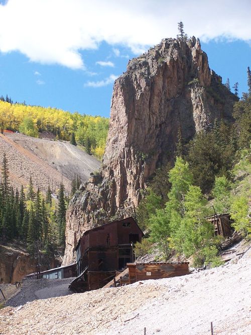 Abandoned mining structure at the base of a rocky cliff, surrounded by trees and colorful autumn foliage.