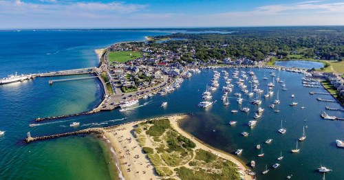 Aerial view of a coastal town with a marina, boats, sandy beach, and lush green areas under a clear blue sky.