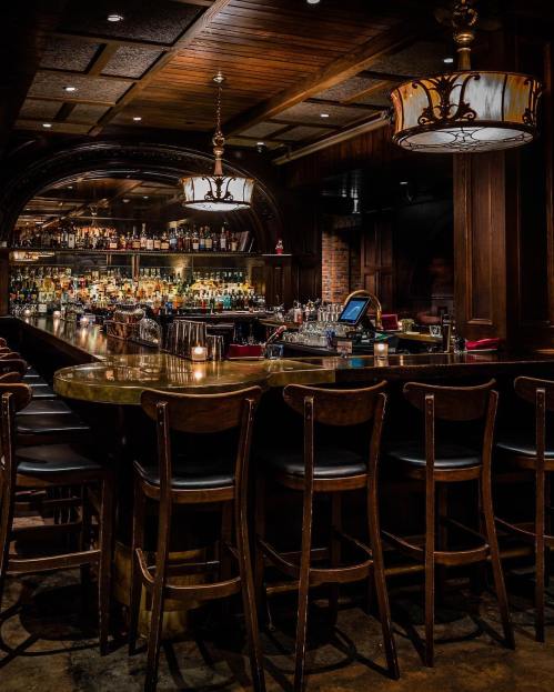 A dimly lit bar with a polished counter, wooden stools, and shelves lined with various bottles in the background.