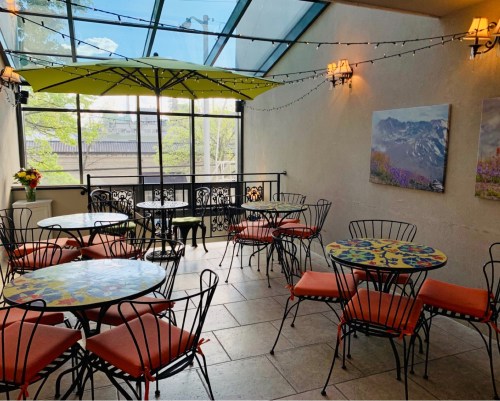 Bright indoor café space with colorful tables, chairs, and large windows, featuring an umbrella and decorative lighting.