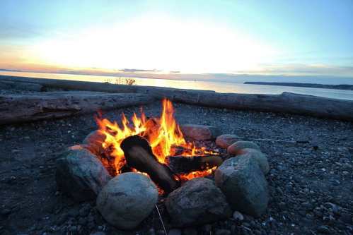 A campfire surrounded by stones, with a serene sunset over the water in the background.
