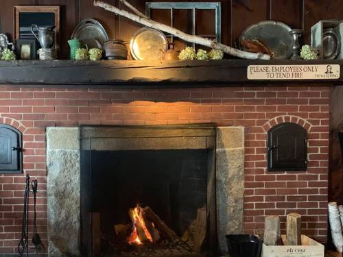 A rustic fireplace with a brick surround, wooden mantel, and decorative items above, including a sign and greenery.