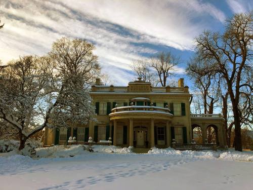 A grand, snow-covered mansion with green shutters, surrounded by bare trees and a blue sky with wispy clouds.