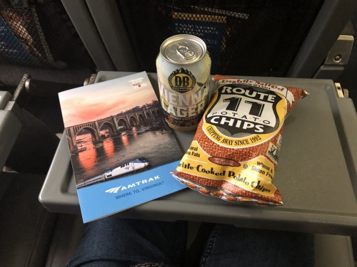 A can of beer, a bag of potato chips, and an Amtrak brochure on a tray table in an airplane.