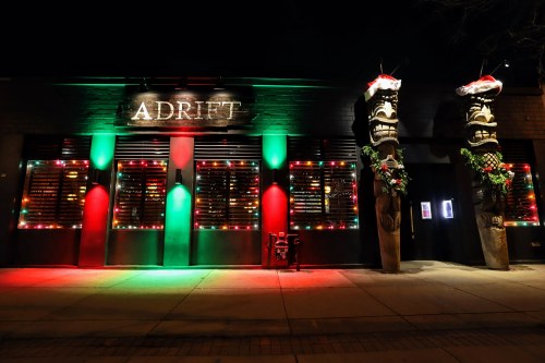 A brightly lit bar named "Adrift" with festive decorations and tiki statues, illuminated in red and green lights at night.