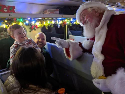 A joyful Santa interacts with a smiling child on a decorated train, while an adult looks on happily.