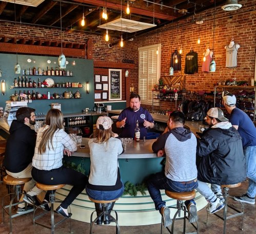 A group of people seated at a bar, listening to a bartender in a rustic setting with exposed brick and warm lighting.