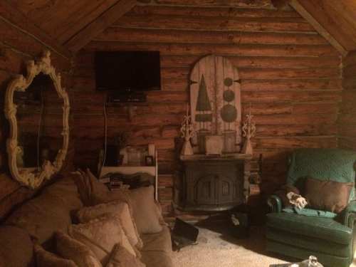 Cozy log cabin interior with a fireplace, green armchair, mirror, and rustic decor on wooden walls.