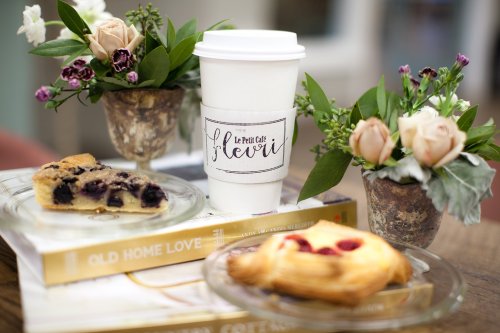 A coffee cup with "Le Petit Café" label, pastries, and small floral arrangements on a table with books.