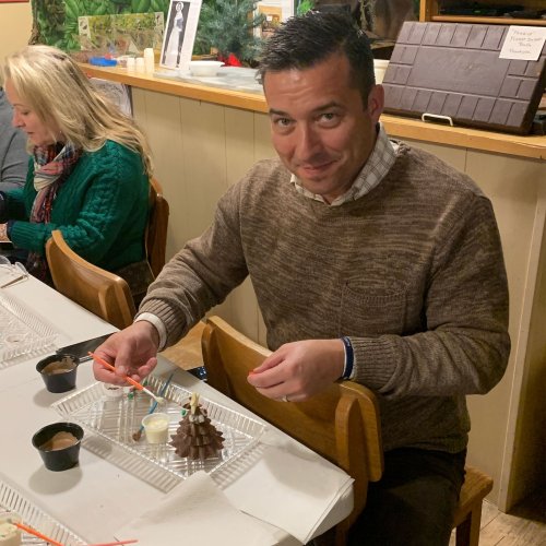 A man smiles while sitting at a table with dessert items, surrounded by people in a cozy setting.